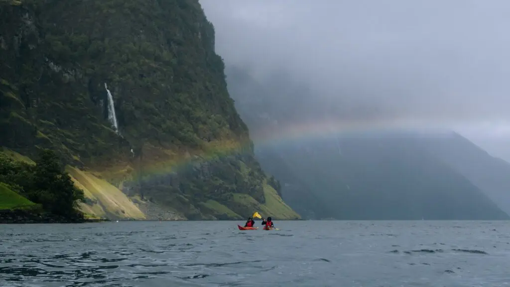 deux personnes faisant du kayak sur un fjord, avec un arc-en-ciel et une cascade