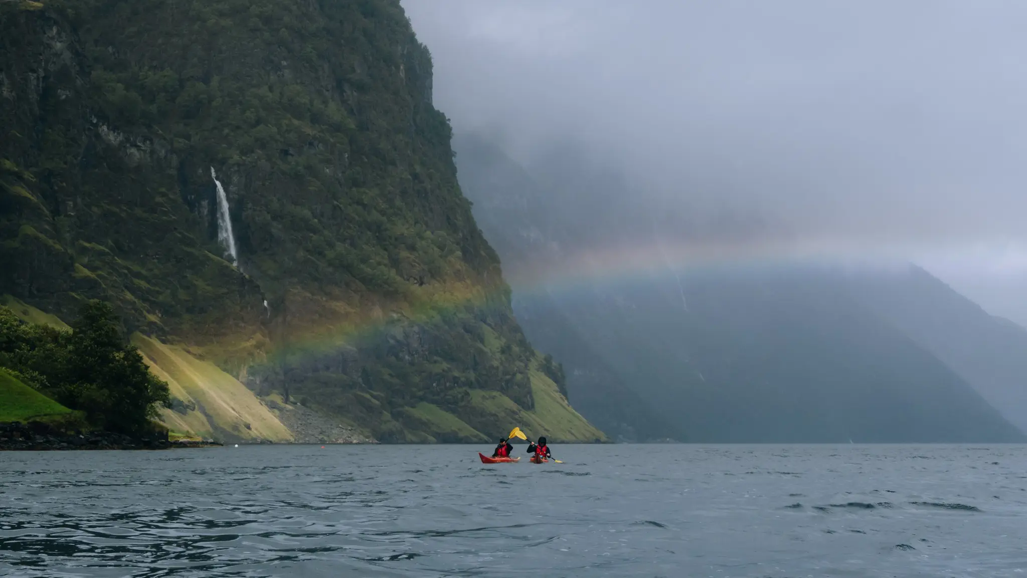 deux personnes faisant du kayak sur un fjord, avec un arc-en-ciel et une cascade