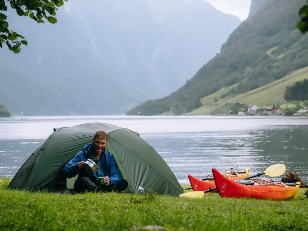 homme versant du café devant une tente au bord d’un fjord avec des kayaks