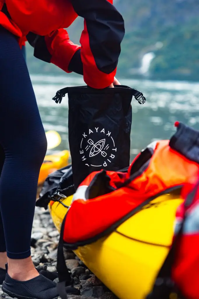 personne préparant un sac étanche à côté d’un kayak sur une rive rocheuse